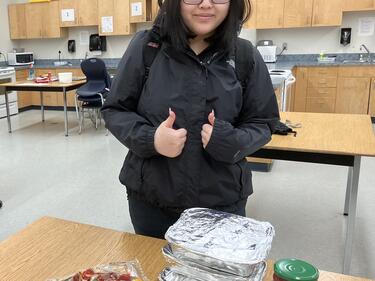 Grade 12 female student with their thumbs up, standing in front of food supplies