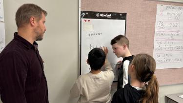 Three students working on math program hung on wall while teachers observes.